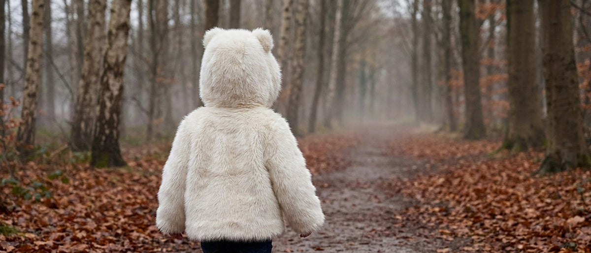 A toddler walking away down a misty autumn woodland path in the Snowfall Little Bear faux fur coat, hood up with bear ears in silhouette