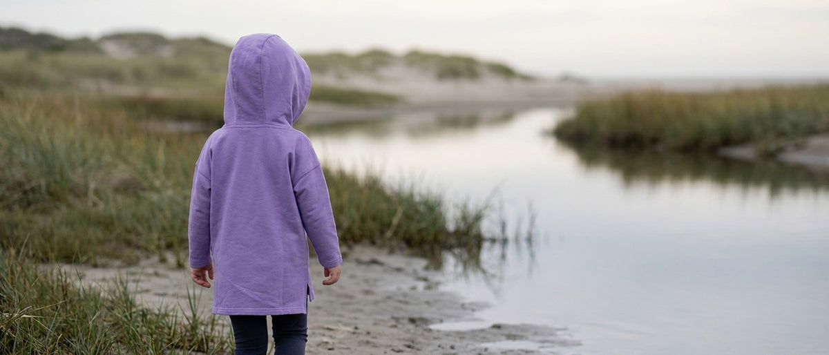 A child walking away along a tidal creek in the Story-Keeper lavender hooded tunic, hood up, longer back hem visible